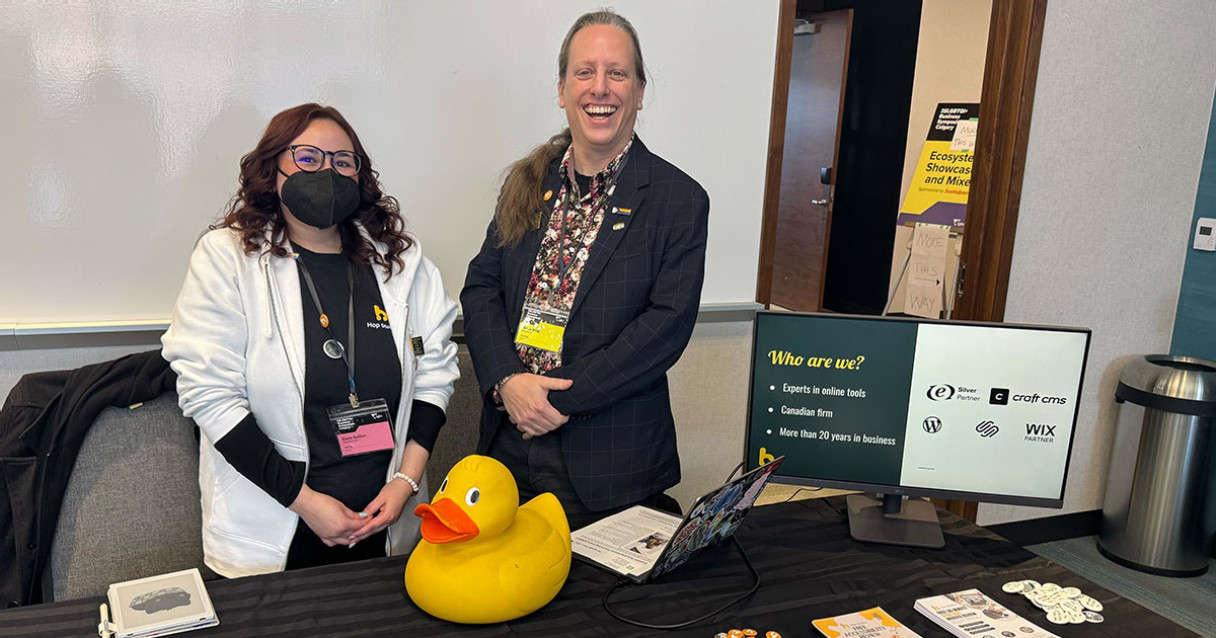A woman in a mask and a man laughing stand behind a table with a monitor and a giant rubber duck on it