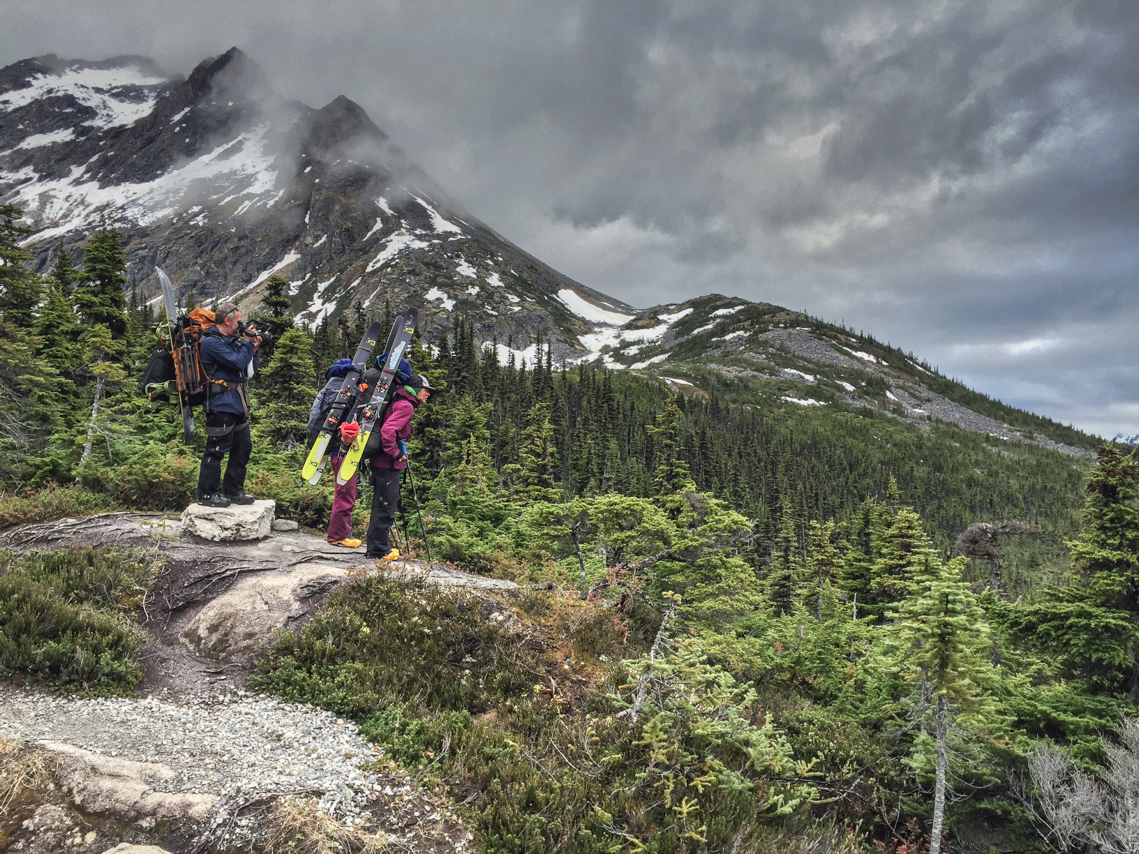 Two hikers loaded with backpacks and skis survey a mountain view from a rocky outcrop